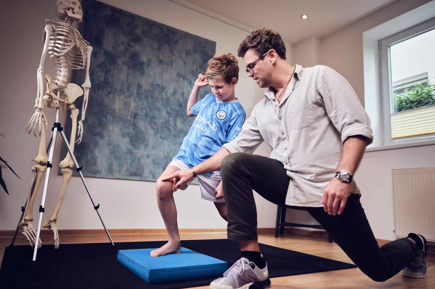 Trainingstherapie mit einem Kind: Der Junge balanciert auf einem Therapiekissen, der Behandler führt sein Knie