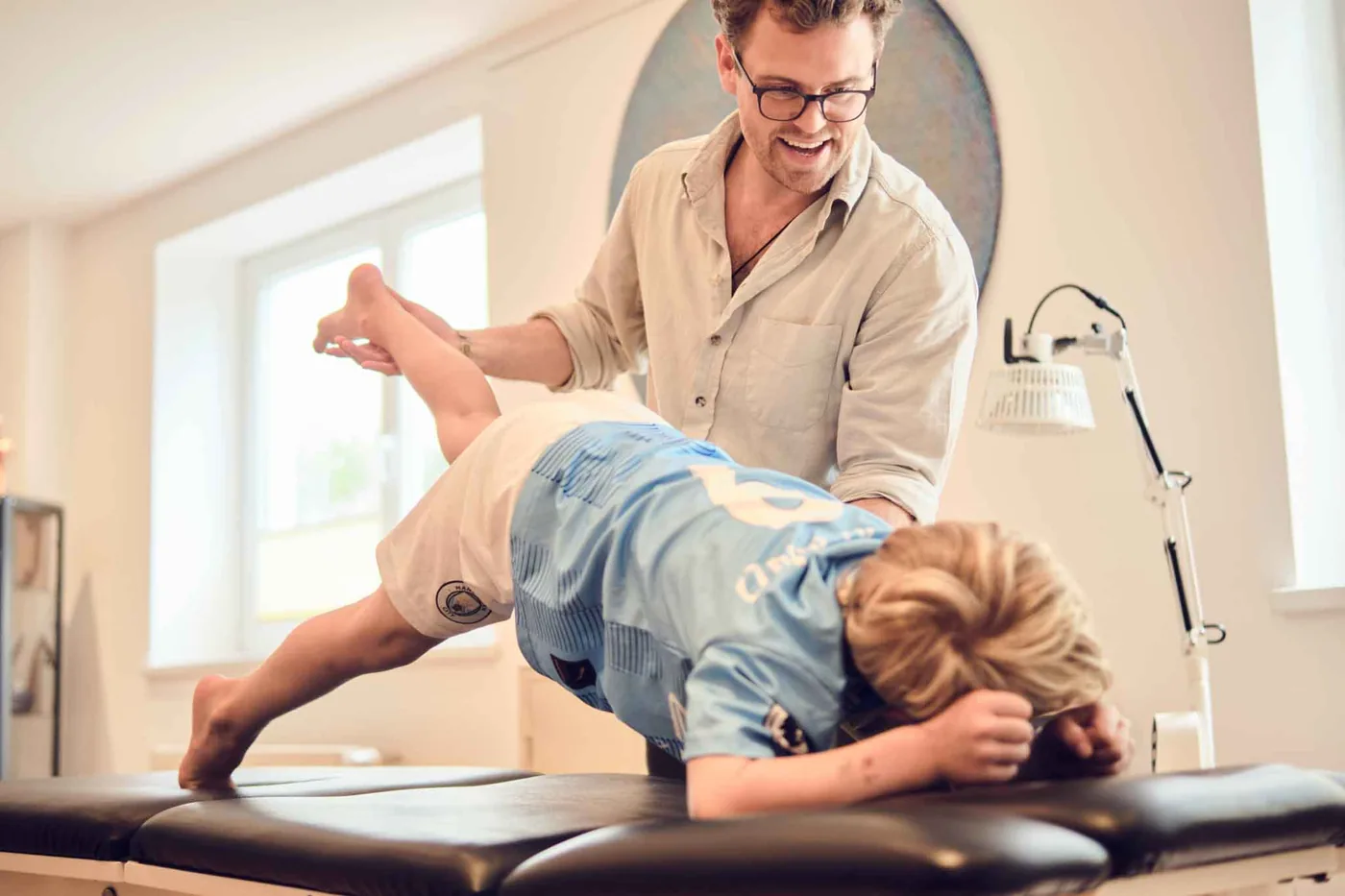 Kräftigungsübung auf der Behandlungsliege: Der Behandler stützt das angehobene Bein eines Jungen