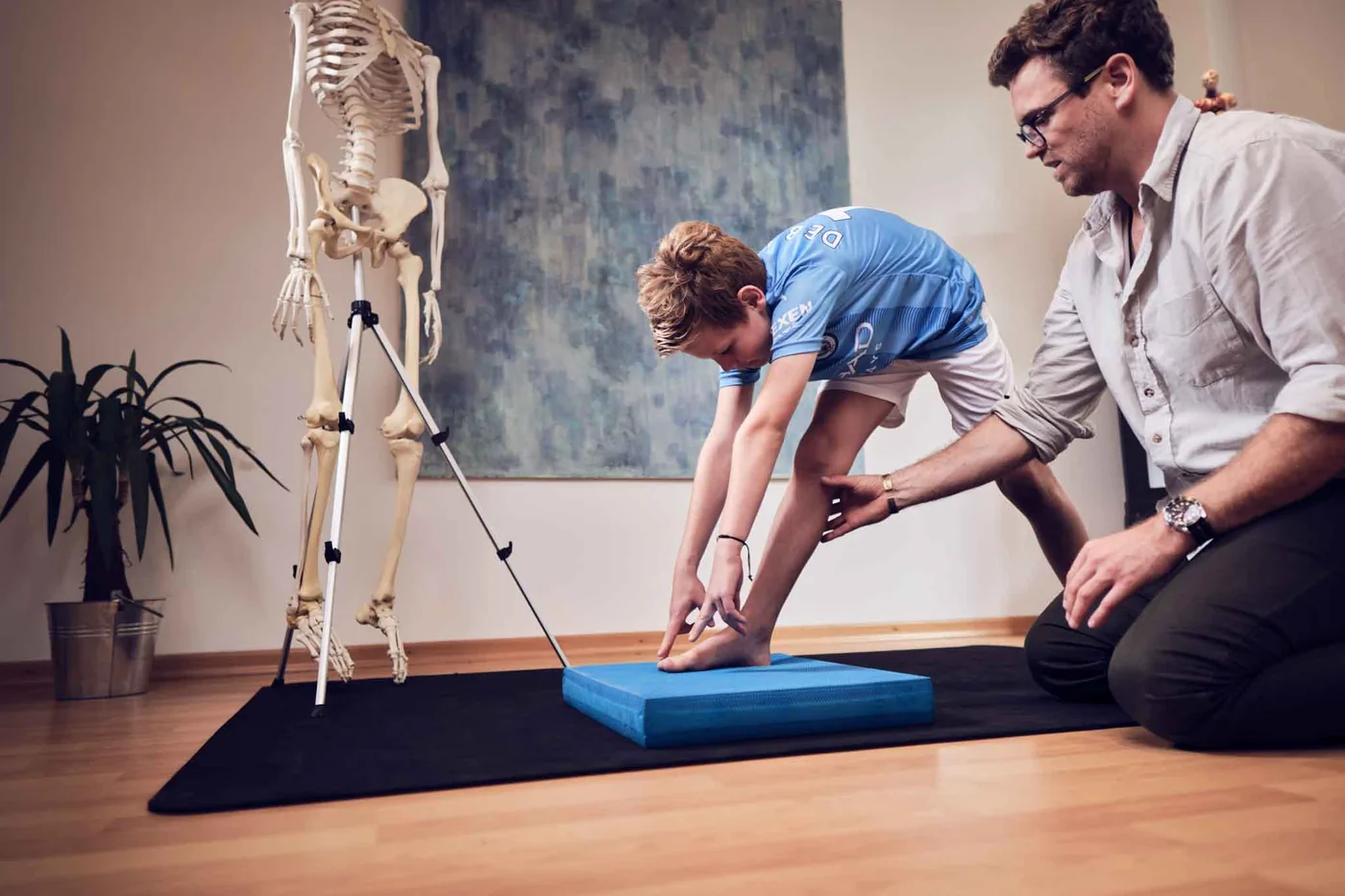 Beweglichkeitsübung: Der Junge beugt sich auf dem Balancekissen nach vorn, der Behandler sichert sein Knie